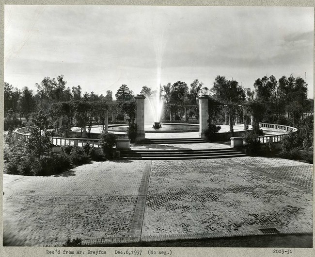 Black and white photograph of circular fountain surrounded by tall trees and stone paths leading up to it. Enclosing the fountain are Romanesque  columns.