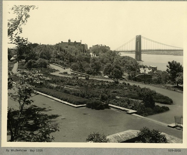 Black and white photograph of multi-leveled park, with dirt paths winding around large green patches of land. In the distance are buildings, a body of water, and a cable bridge going across it.