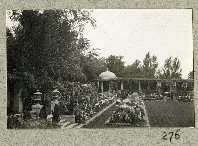 Black and white photograph of garden. There is an open green rectangle in the center, with other smaller rectangles inside holding different plants and flowers. There are stone steps leading to a column of roman style pillars, and an open dome.