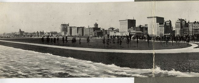 Black and white photograph of large oval area with a dirt path around a large central oval covered in grass and planted with trees. Waves of water are coming up from one side, with another lined with buildings