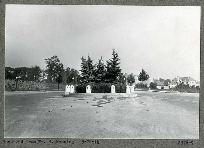 Black and white photograph of dirt lot with circular green space at the center. The center of that space is planted with large trees and bushes.