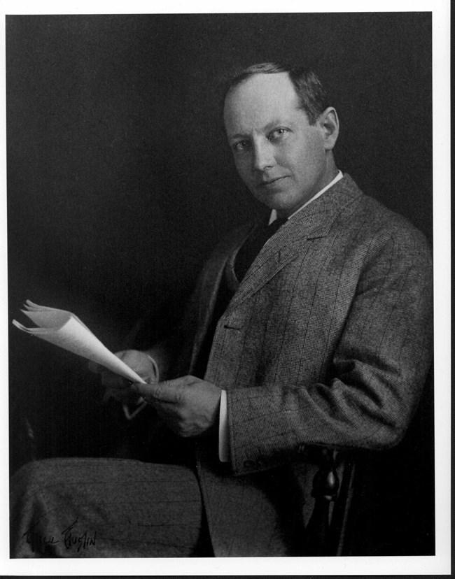 Black and white photograph of man seated at chair holding a piece of paper in a suit and tie