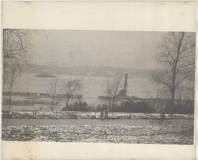 Black and white photograph of pathway lined with grass and benches looks out over a body of water. The few trees on land have no leaves, and there is snow on the ground.