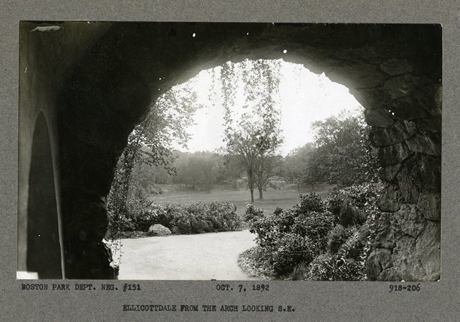Black and white photograph of view under stone archway looking out onto open green space. The dirt path out of the tunnel is lined with bushes, and in the distance are few scattered trees.