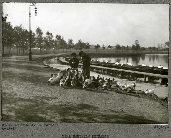 Black and white of two young boys standing by body of water with small boats on it feeding a bunch of ducks