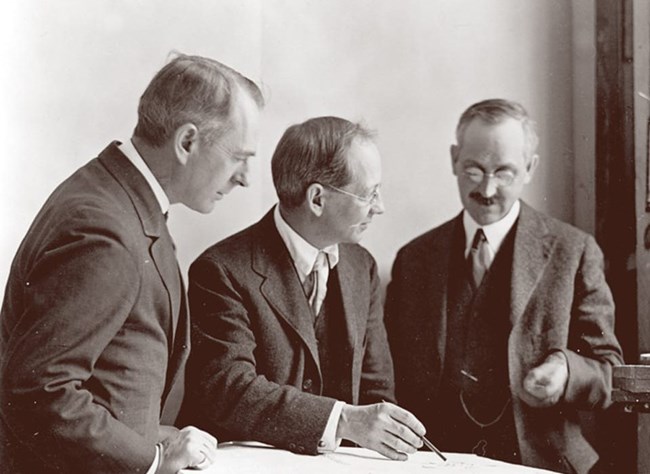 Three men in suits talk around table with paper on it