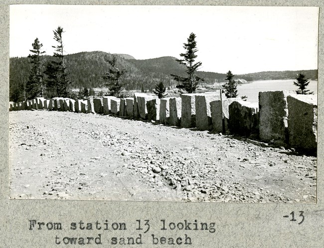 Black and white of dirt road with stone wall and on the other side is open space, and a mountain rising with trees on it and in the distance