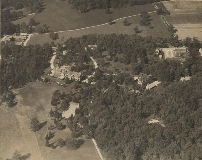 Black and white aerial of large home with two roads leading to it, withfront and surroundings densely planted, but an open yard next to the house.