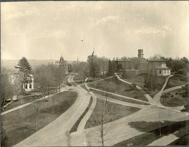 Black and white aerial of grassy, slightly hilly area with many paths going across, with buildings on the edge and some trees spread out.