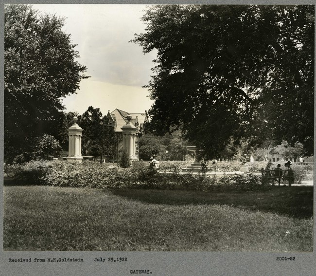 Black and white photograph of green space with arches, benches, trees and people