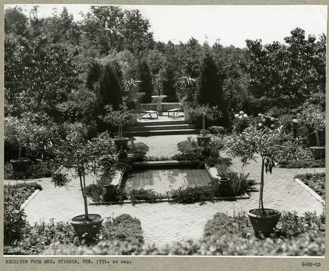 Black and white of densely planted garden with stone paths in a square shape with a square pool in the center and a seating area in the distance up a few stairs.