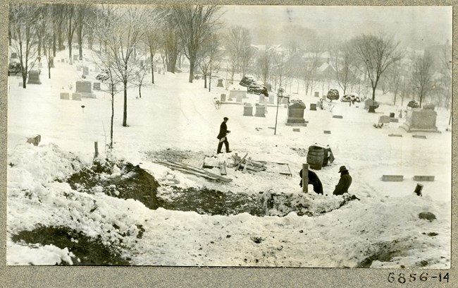 Black and white photograph of cemetery with graves spread out and snow on the ground. There are some people in coats moving snow with shovels. On the edge of the cemetery is a row of trees, and then homes.