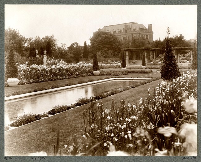 Black and white of flat rectangular garden with plantings on the edge, grass and then a rectangular pool. In the distance a house rises in the hill.