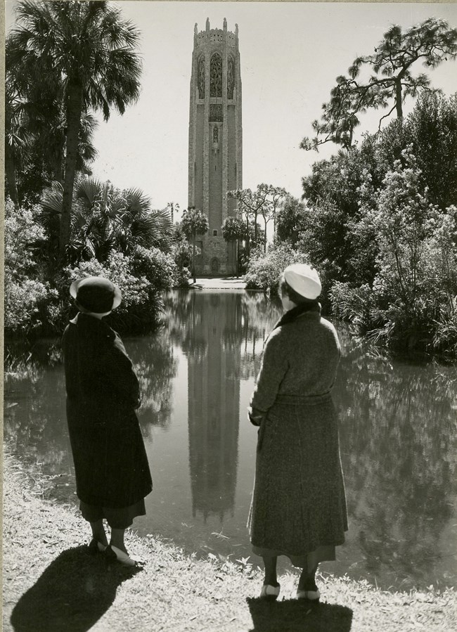 Two women stand in front of body of water with trees around edges and tower at the opposite end.