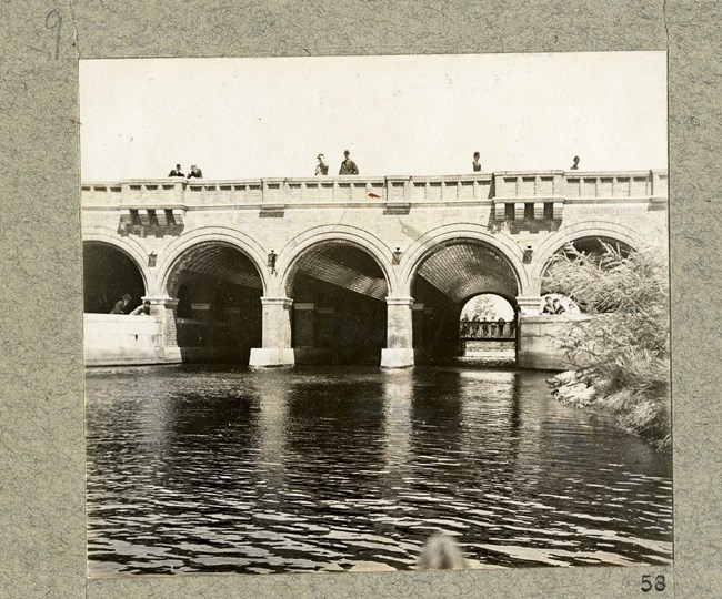 Black and white of stone bridges with arches over a body of water with people on top of the bridge.