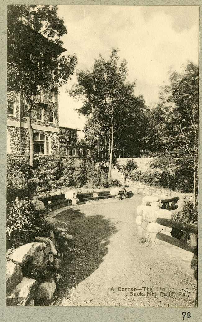 Black and white photograph of dirt path in front of a large stone home. The path is lined by a stone wall, that occasionally breaks for benches. Right by the path are shrubs, and closer to the house are larger trees.