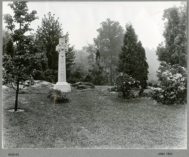 Black and white photograph of intricate, white stone cross on grass, surrounded by different trees and bushes.