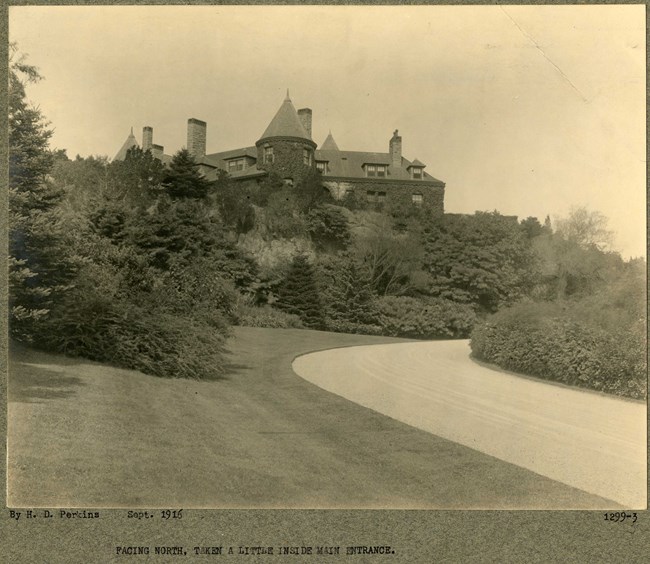 Black and white of dirt road leading up to large home on a hill with grassy area on one side, then dense plantings, and more shrubs on the other side.