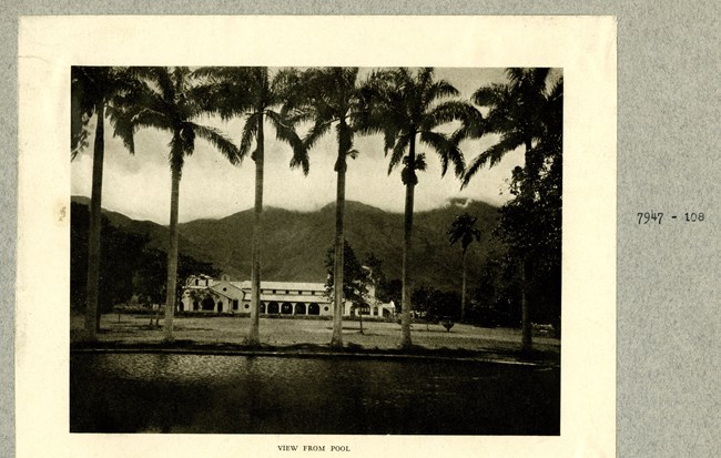 Black and white photograph of country club perched at the bottom of towering green hills. The picture is taken from the pool, which is in the front, followed by a line of palm trees, open grassy area, the building, and then the mountains.