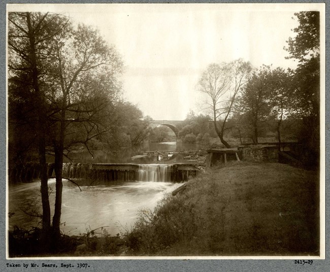 Black and white of small waterfall. Upstream the water is flat and there is a large bridge over the water.