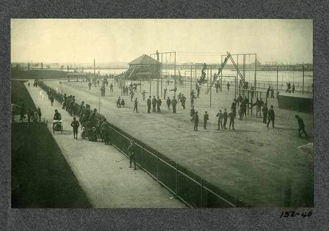 Black and white of large dirt area with playground equipment like ladders and jungle gyms and many men in suits standing and climbing on structures. Walking path on one side, body of water on the other.