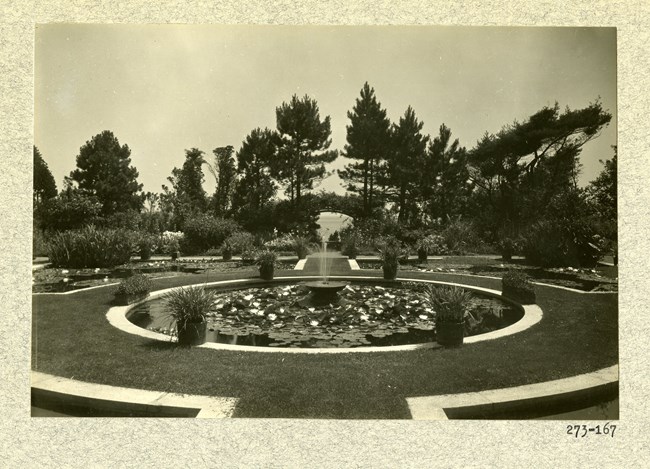 Black and white of symmetrical garden with fountain in middle and trees and plants, with water in the far distance.