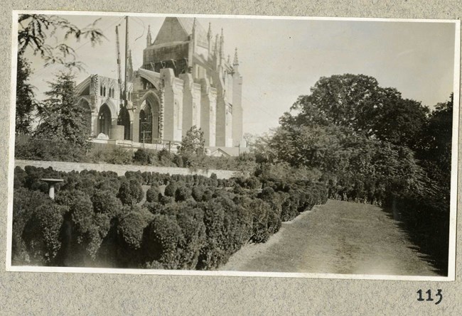 Black and white photograph of white building in distance being built, while in front there are lines of plantings