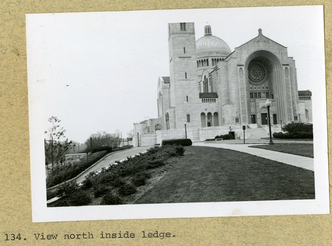 Black and white photograph of large building with dome on top and large intricate arch in the middle. The grounds in front of the church are well manicured with bushes along the road.