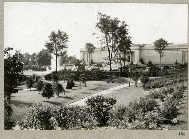 Black and white photograph of white stone building behind a large open green space. The green space has paths that cut through it, which are lined with plants.
