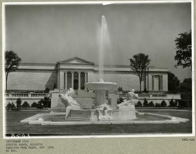 Black and white photograph of large stone fountain with three angles around the fountain. Behind the fountain is a large white building with stone columns
