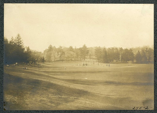 Black and white of bottom of hill with large grassy area and trees on the edges. People are siting on the grass, some look to be playing baseball