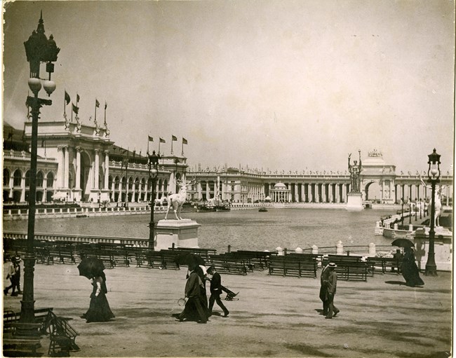 People walk along edge of water with palatial white large buildings rising around the water.