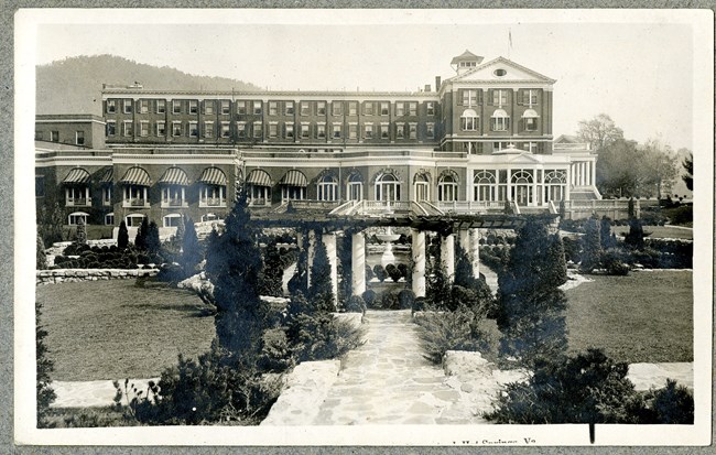 Black and white photograph of large building with many windows with garden in front. Garden has a stone path going to a fountain, which is surrounded by plantings, with some grassy space.