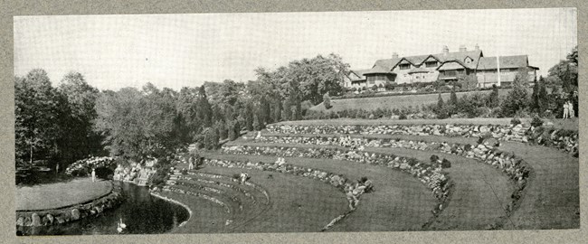 Black and white of large house on top of hill with grassy hill leading down, and becoming terraces, separated by rocks, like a stage. At bottom is water moat, then flat area of grass. There are people scattered on the steps.