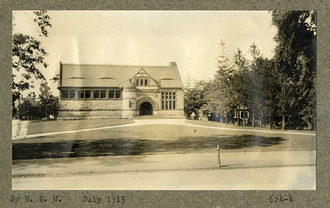 Black and white of large grassy area with two diagonal paths leading to a stone building. Trees line the edge of the property.