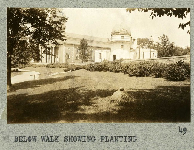 Black and white of small grassy hill with a path at the bottom and stairs leading up to a line of shrubs, a stone wall, and then a large stone building.