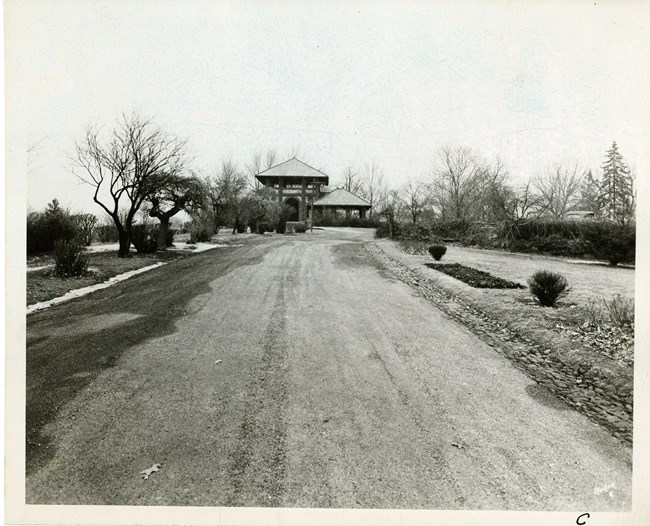 Black and white of dirt road lined with grassy area with some trees without leaves, and a stone structure at the end of the path.