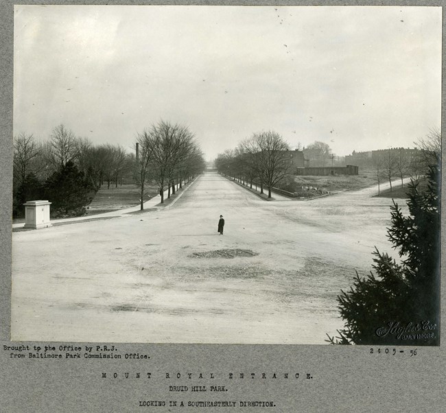 Black and white of dirt road lined with trees and a pathway with grassy area on the outside. A man is standing in the middle of the road.