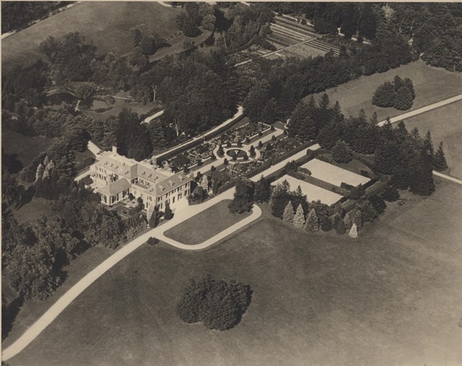 Black and white aerial photograph of large estate, with a large garden in front of an even larger home. The garden has many different planting areas, and a central area in the middle. Around the house are more areas of green