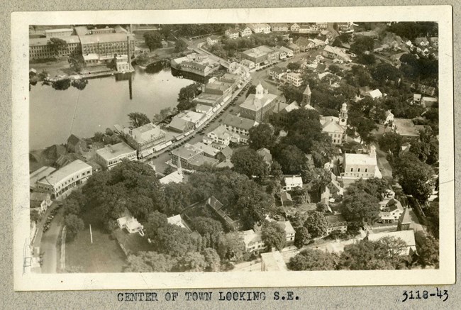 Black and white aerial of school campus next to river, with city buildings right outside dense greenery of campus.