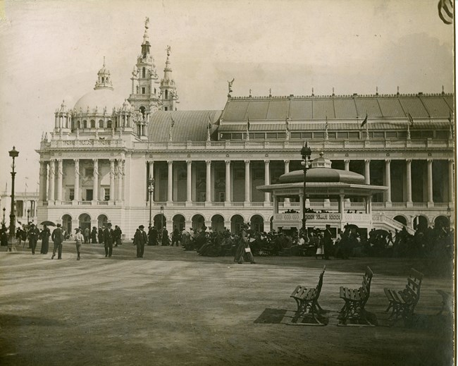 Black and white photograph of large white building with Roman like columns. In front of the building is a large group of people, some sitting, some walking.