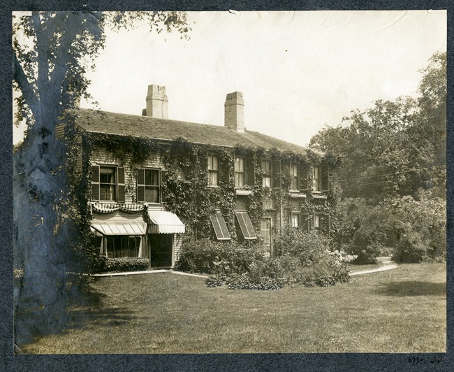 Black and white of home covered with vines on flat piece of grass with trees planted at the edge, and a dirt path following closely around the house.