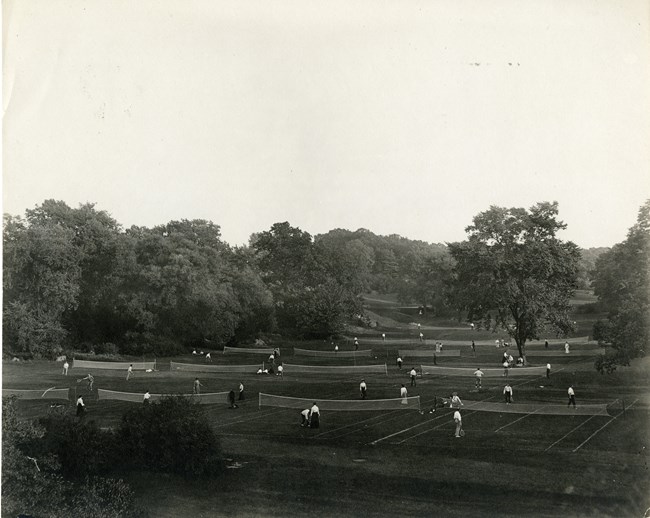 Black and white open field with many trees around with many tennis courts and players in the center.