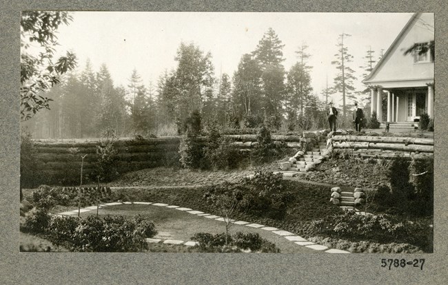 Black and white of sunken garden surrounded by wall made of fallen tree logs with two men standing on stairs at top of garden with building behind them.