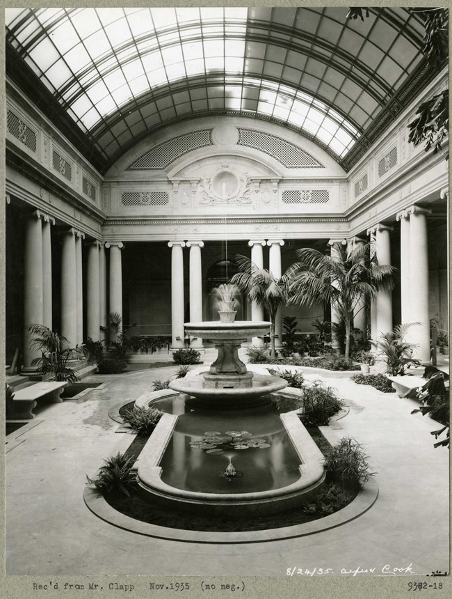 Black and white photograph of indoor garden area with glass tile ceiling. The centerpiece is a fountain, the room is supported by large white pillars and there are plantings scattered across the room
