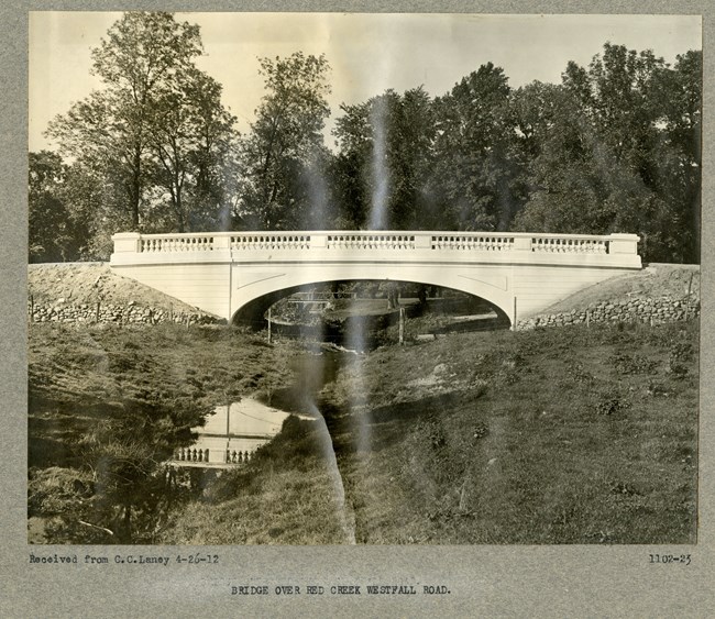 Black and white of small white stone bridge over small creek with grass on the side and trees behind the bridge.