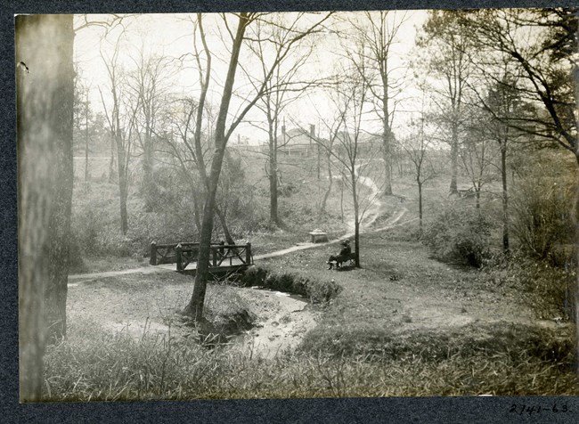 Black and white of path going up a hill with a wooden bridge , bench with people sitting on it, and buildings up the mountain.