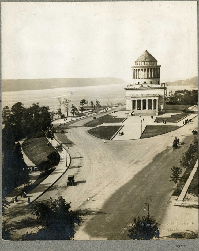 Black and white aerial view of road leading up to large white building with pillars and stairs, with park space in front and around the building.