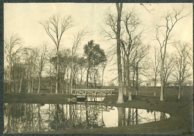 Black and white of small body of water surrounded by grass area with trees. By the water is a bridge, and all the trees are reflected in the water.