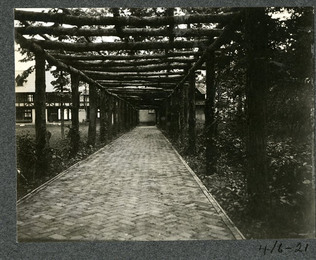 Black and white of stone path next to grassy area with a wood log roof above and a building at the end of the path.